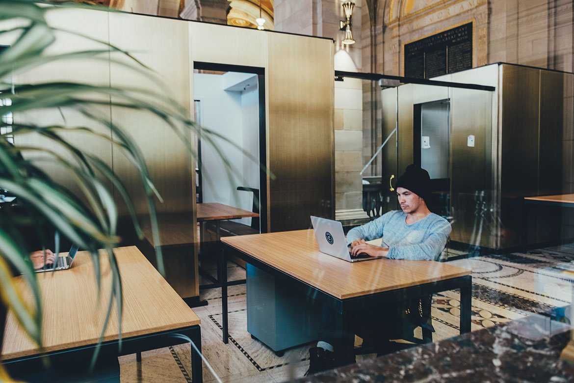 A man in a beanie working on a laptop at a desk in an open office space